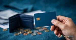 Hand holding a plain dark blue credit card with a gold chip, with an open wallet and scattered coins on a blue surface behind it